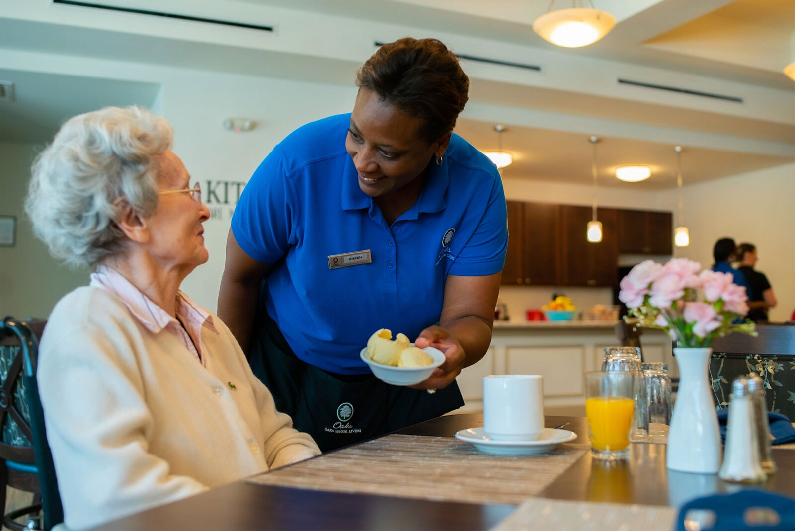 Caregiver serving food to smiling elderly woman in dining area.