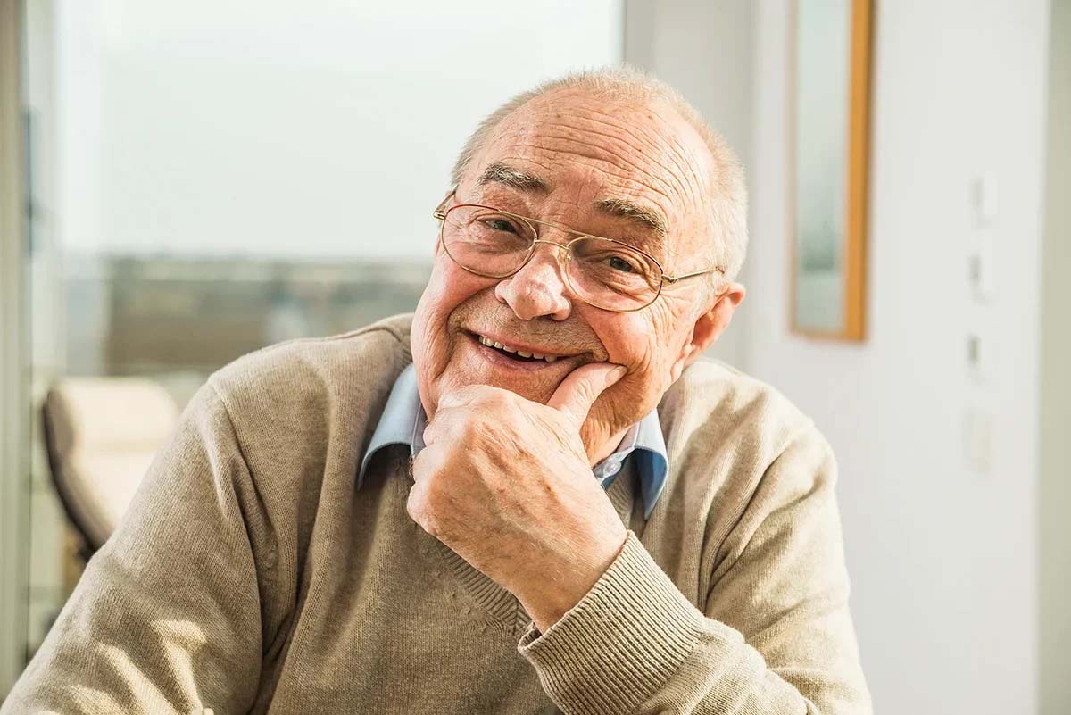 Smiling elderly man with glasses, resting chin on hand.